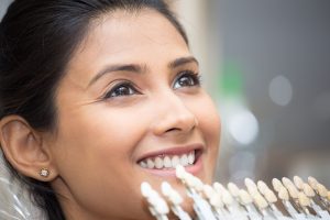 Close-up of smiling woman with white teeth; dental shade guide held near her mouth for color comparison